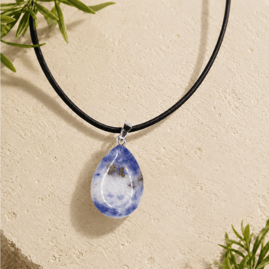 Blue and white stone pendant shaped into a teardrop on a black faux fur leather cord laying on a cream coloured stone slab and green leaves on the sides. 