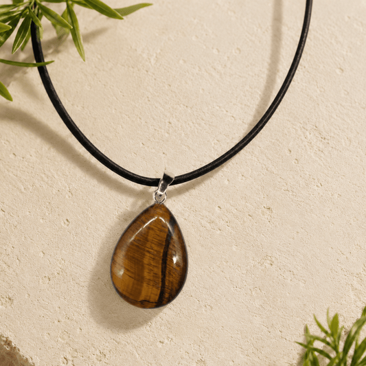 A teardrop-shaped pendant necklace made from polished tiger's eye stone with a black cord and silver-toned metal clasp, displayed against a textured beige wall with small green leaves in the corners.