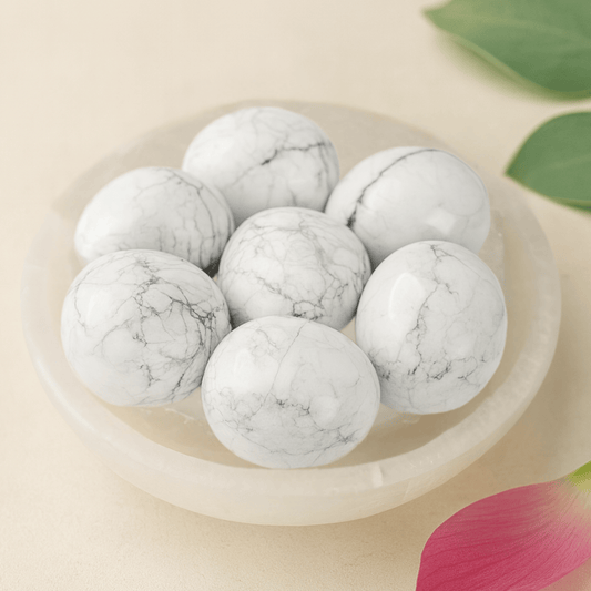 Seven White Howlite tumbled stones in a selenite bowl with a beige background and a soft pick petal and green leaf.
