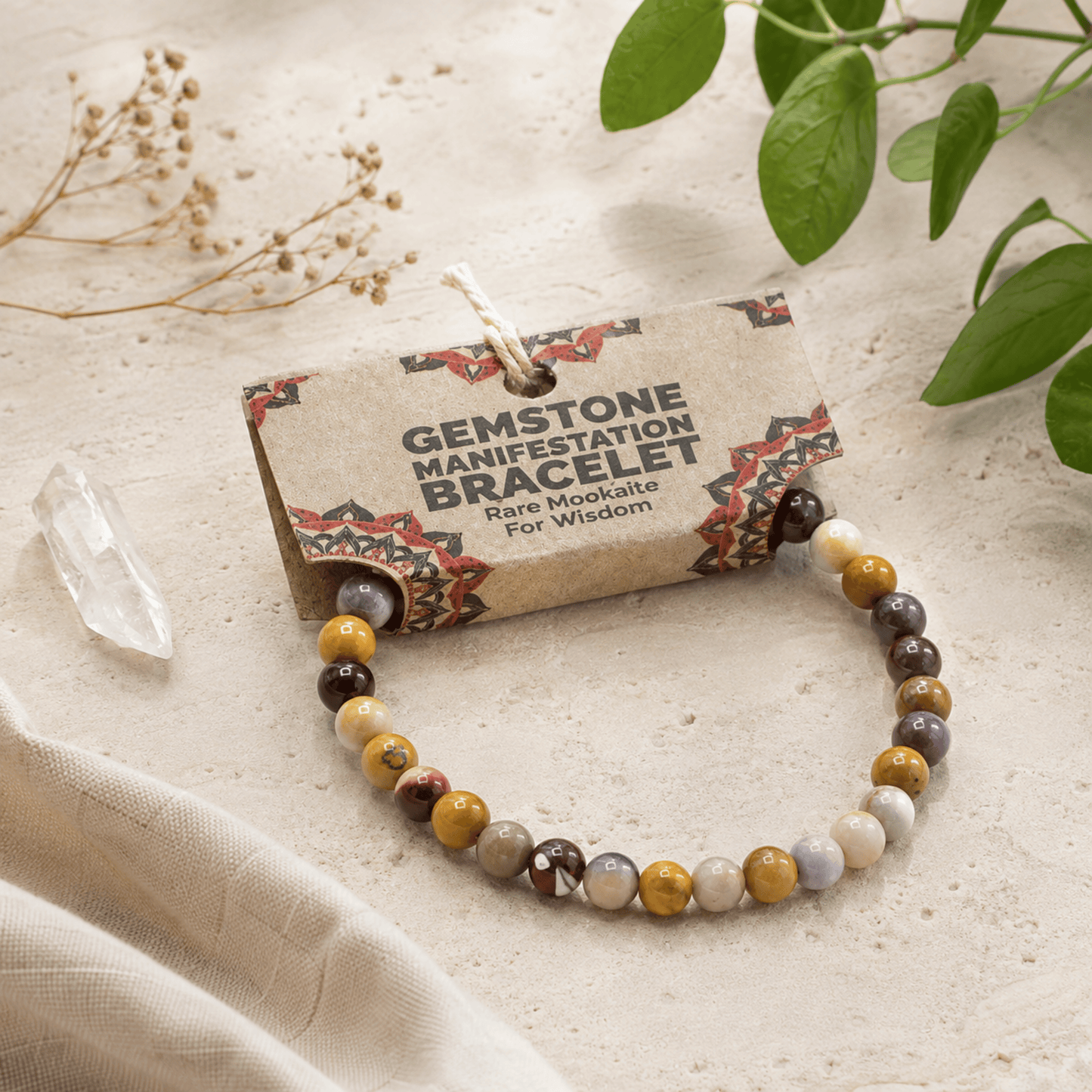 A gemstone manifestation bracelet with multi-colored polished beads, displayed on its kraft paper tag which reads 'GEMSTONE MANIFESTATION BRACELET Rare Mookate For Wisdom'. The scene is styled with a clear crystal point, dried flowers, and green leaves on a textured beige fabric background.