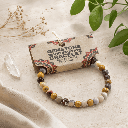 A gemstone manifestation bracelet with multi-colored polished beads, displayed on its kraft paper tag which reads 'GEMSTONE MANIFESTATION BRACELET Rare Mookate For Wisdom'. The scene is styled with a clear crystal point, dried flowers, and green leaves on a textured beige fabric background.
